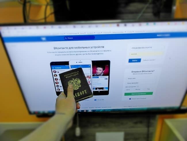 Рука человека, протягивающего российский паспорт к монитору, в котором видна открытая страница «ВКонтакте».